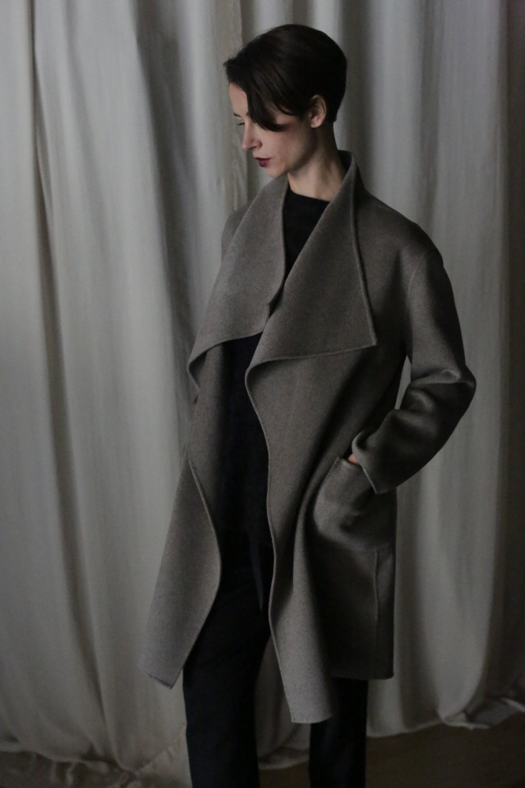 Wearing the Winston Long Double-face coat in undyed natural, a person with short hair looks down, hands in pockets over dark clothing—a true wardrobe foundation against soft, light-colored fabric curtains.