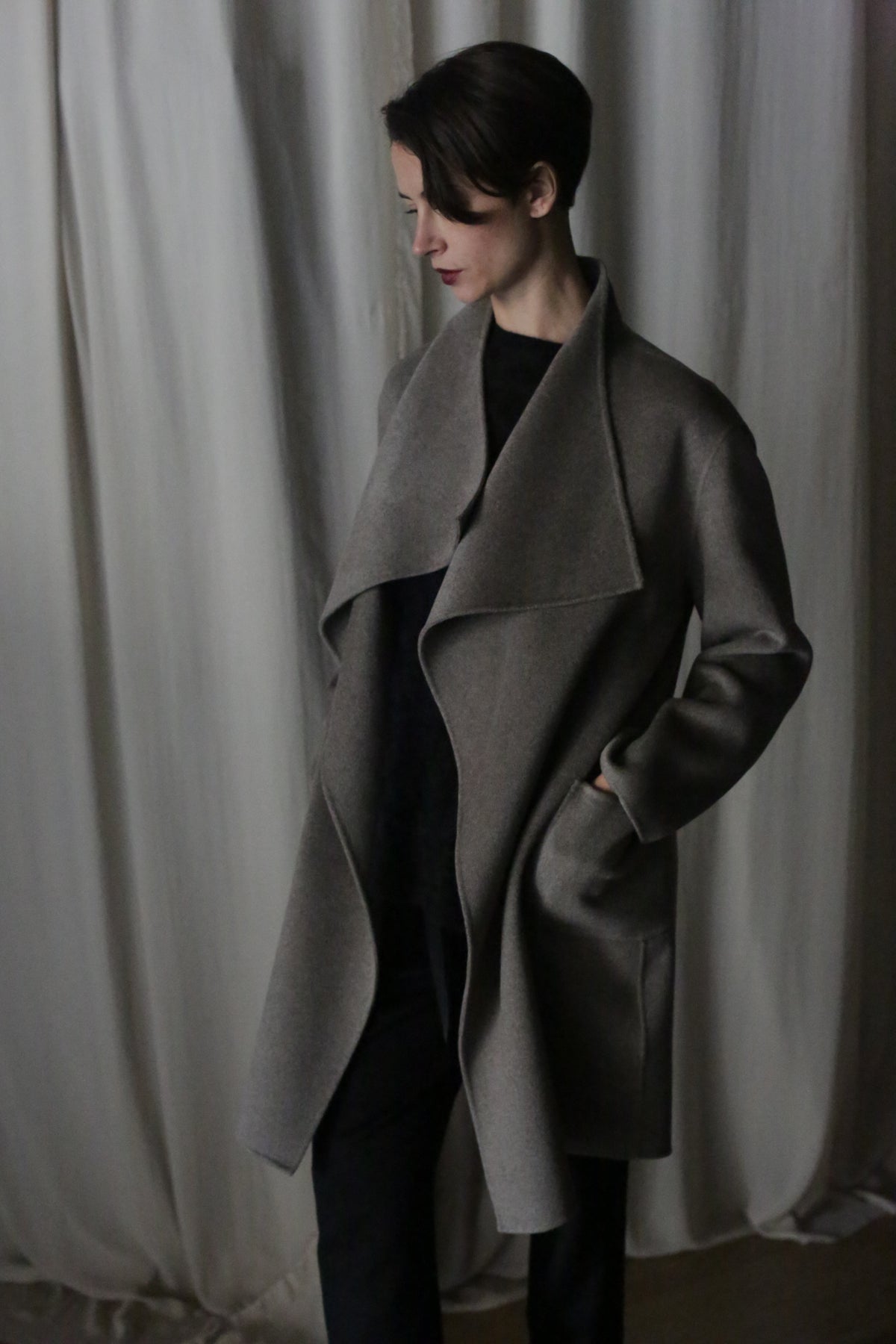 Wearing the Winston Long Double-face coat in undyed natural, a person with short hair looks down, hands in pockets over dark clothing—a true wardrobe foundation against soft, light-colored fabric curtains.