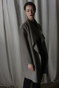 A person stands before a curtain in the Winston Long Double-face | Undyed Natural, a perfect wardrobe foundation. Dim lighting casts shadows as they gaze slightly downward with short hair, embodying timeless neutrals.