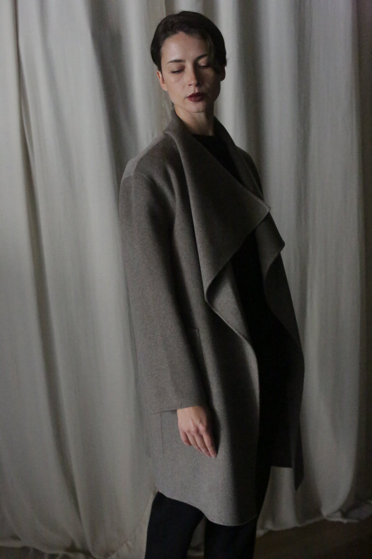 A person stands before a curtain in the Winston Long Double-face | Undyed Natural, a perfect wardrobe foundation. Dim lighting casts shadows as they gaze slightly downward with short hair, embodying timeless neutrals.