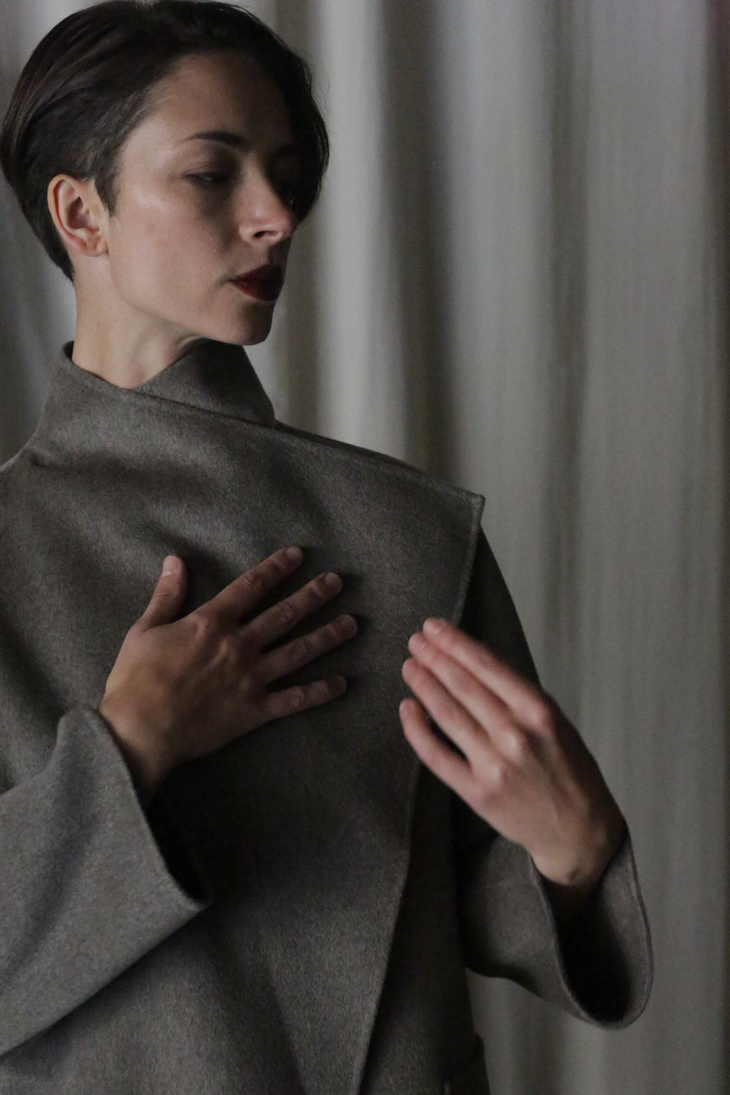 A person with short hair, wearing the Winston Long Double-face in Undyed Natural, stands against a neutral backdrop. Their left hand rests on their chest as their right is raised. Soft lighting casts subtle shadows on their face, showcasing this wardrobe foundation piece.