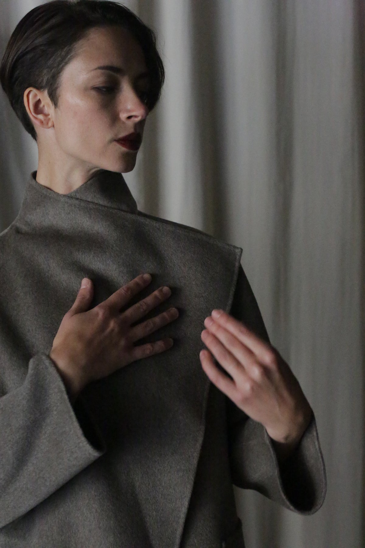 A person with short hair, wearing the Winston Long Double-face in Undyed Natural, stands against a neutral backdrop. Their left hand rests on their chest as their right is raised. Soft lighting casts subtle shadows on their face, showcasing this wardrobe foundation piece.
