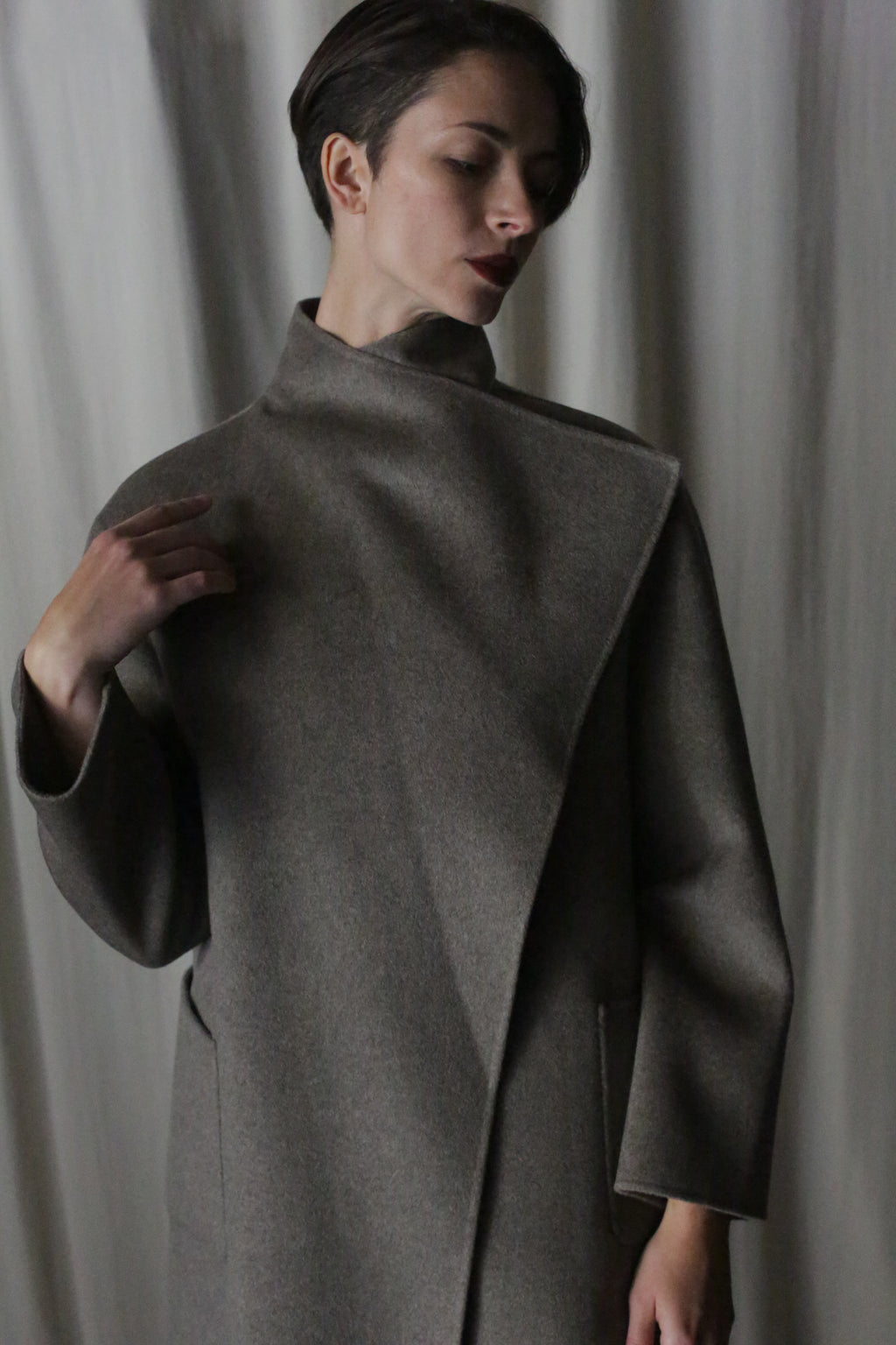 Modeling short hair and the Winston Long Double-face coat in Undyed Natural, an enduring neutral piece suitable for any wardrobe, they gently touch the high collar against a softly lit backdrop, exuding a pensive elegance.