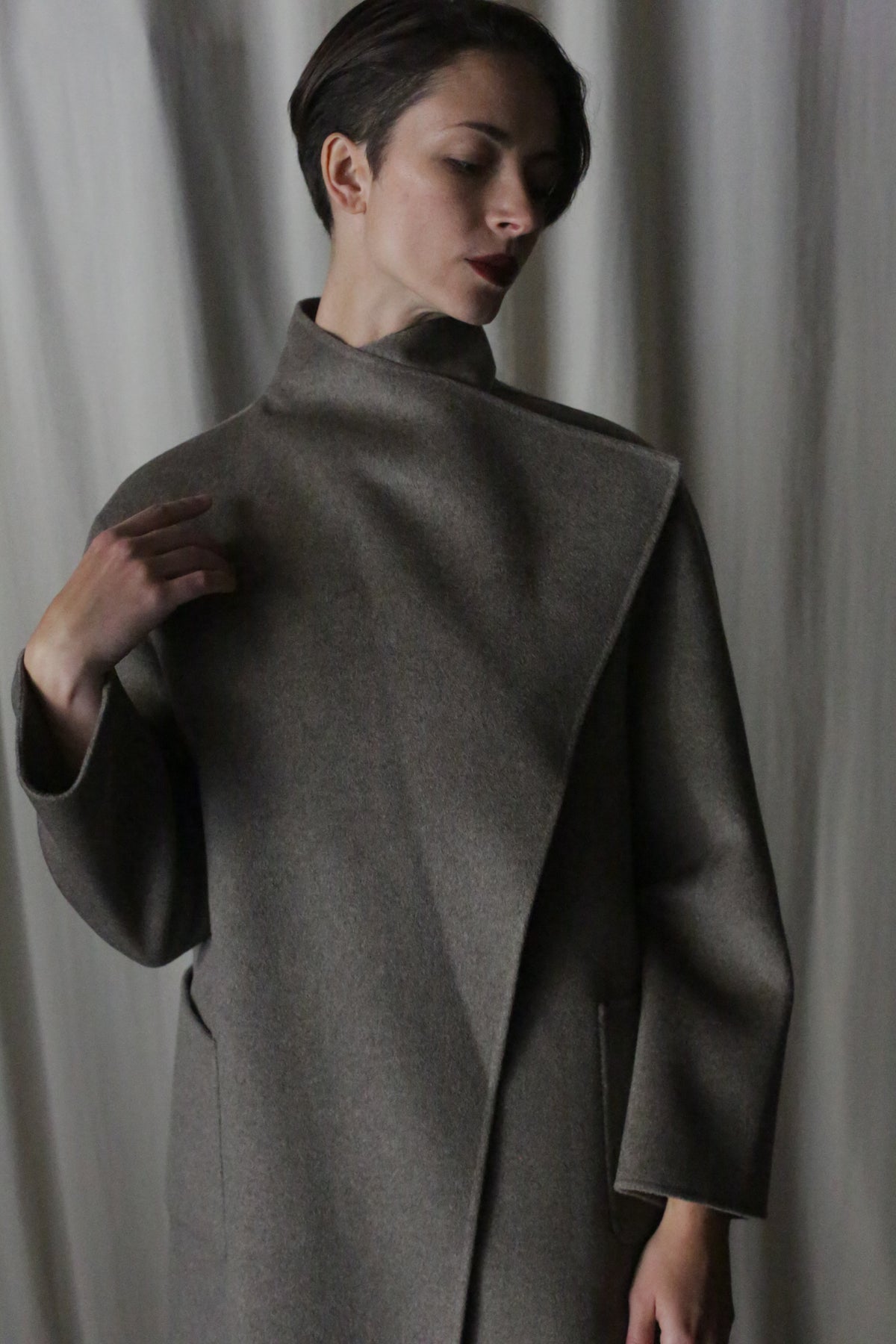 Modeling short hair and the Winston Long Double-face coat in Undyed Natural, an enduring neutral piece suitable for any wardrobe, they gently touch the high collar against a softly lit backdrop, exuding a pensive elegance.