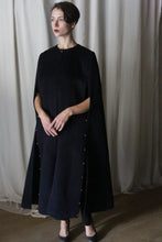 Load image into Gallery viewer, A person stands in front of light-colored curtains, wearing the Opera Cape by Atelier Lan Jaenicke, a long, dark wool cape-like garment adorned with gold buttons along the sides. Crafted from Suri alpaca, they have short dark hair and are looking slightly to the side, with arms gently crossed.