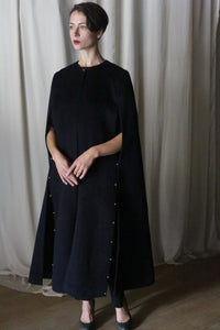 A person stands in front of light-colored curtains, wearing the Opera Cape by Atelier Lan Jaenicke, a long, dark wool cape-like garment adorned with gold buttons along the sides. Crafted from Suri alpaca, they have short dark hair and are looking slightly to the side, with arms gently crossed.