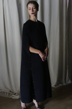 Load image into Gallery viewer, A person wearing an Opera Cape, a long, dark dress-like garment made from soft wool, stands in a dimly lit room with light curtains in the background. They gaze downward with a contemplative expression, evoking a serene and introspective mood reminiscent of an Atelier Lan Jaenicke design.