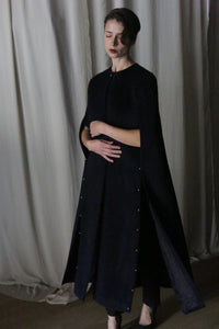 A woman stands against a backdrop of draped fabric, elegantly attired in an Opera Cape made from Suri alpaca wool by Atelier Lan Jaenicke. Her hands are gently clasped, and her eyes are closed, creating a serene and contemplative mood.