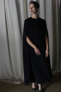 In a dimly lit room, a person stands adorned in the elegant Opera Cape by Atelier Lan Jaenicke. They have short dark hair and are gazing to the side, while draped white curtains shadow their presence.