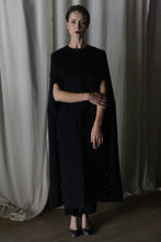 Load image into Gallery viewer, In the dim light, a woman is cloaked in a long, elegant Opera Cape from Atelier Lan Jaenicke, expertly crafted from Suri alpaca wool. She poses in front of draped curtains with one arm crossed over her chest. Her neutral expression is highlighted by dark lipstick.