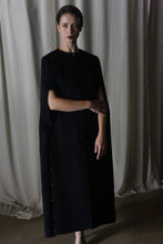 Load image into Gallery viewer, In a dimly lit room, an individual dons a long, dark Opera Cape made from luxurious soft Suri alpaca. Their arms rest gracefully in front of them, while softly draped curtains form the backdrop, evoking a serene and elegant atmosphere characteristic of Atelier Lan Jaenicke&#39;s designs.