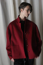 Load image into Gallery viewer, A person with short hair wearing the Bamford Short Double-Face Cashmere coat in wine, designed with a loose fit and buttoned style, stands against a light-colored curtain. Modern fit black clothing beneath complements the ensemble. The individual gazes to the side with a calm expression, embodying the essence of contemporary fashion.