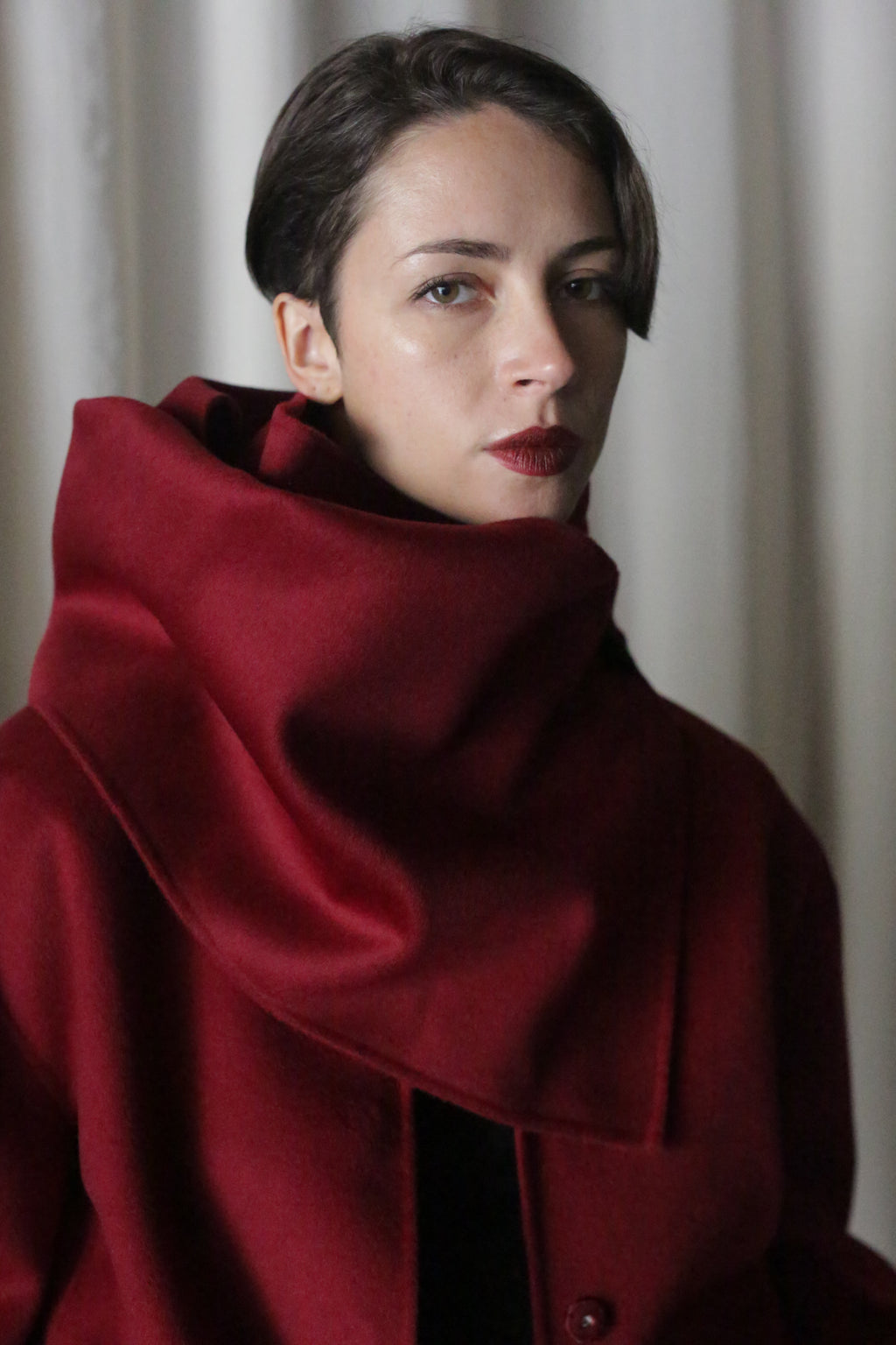 A person with short hair is wearing a rich wine X Scarf, made of double-faced cashmere, featuring a large collar partially covering their neck. They have a neutral expression and stand against a softly lit, neutral background.