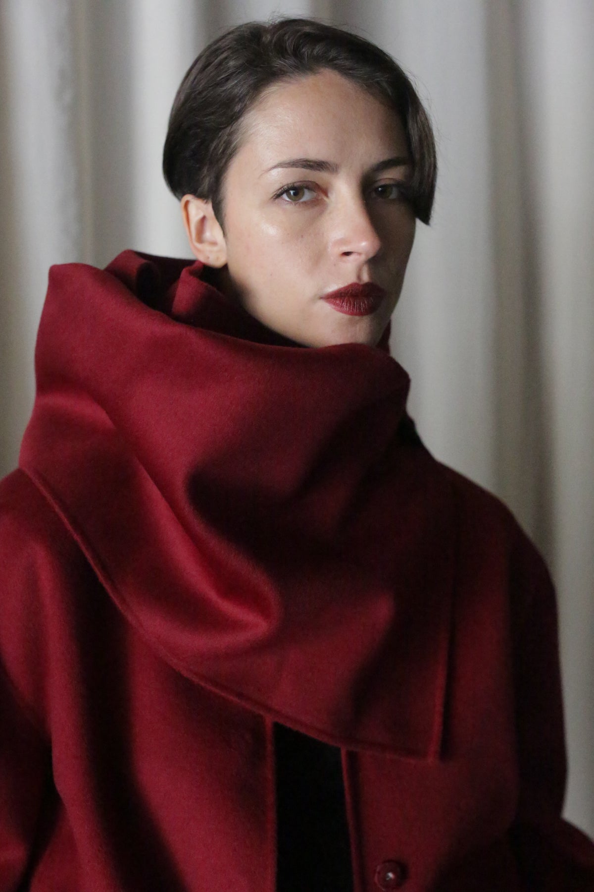 A person with short hair is wearing a rich wine X Scarf, made of double-faced cashmere, featuring a large collar partially covering their neck. They have a neutral expression and stand against a softly lit, neutral background.