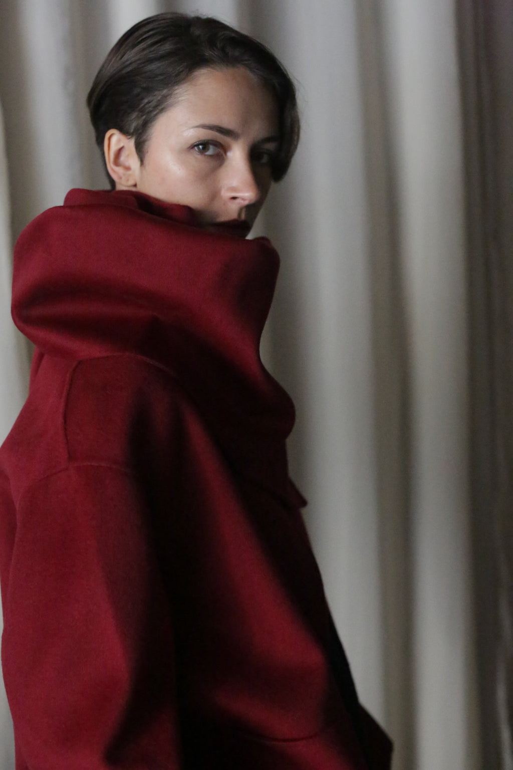Wearing a high-collared wine X Scarf made from double-faced cashmere, a short-haired person glances over their shoulder in a dimly lit room where light curtains subtly outline the scene.