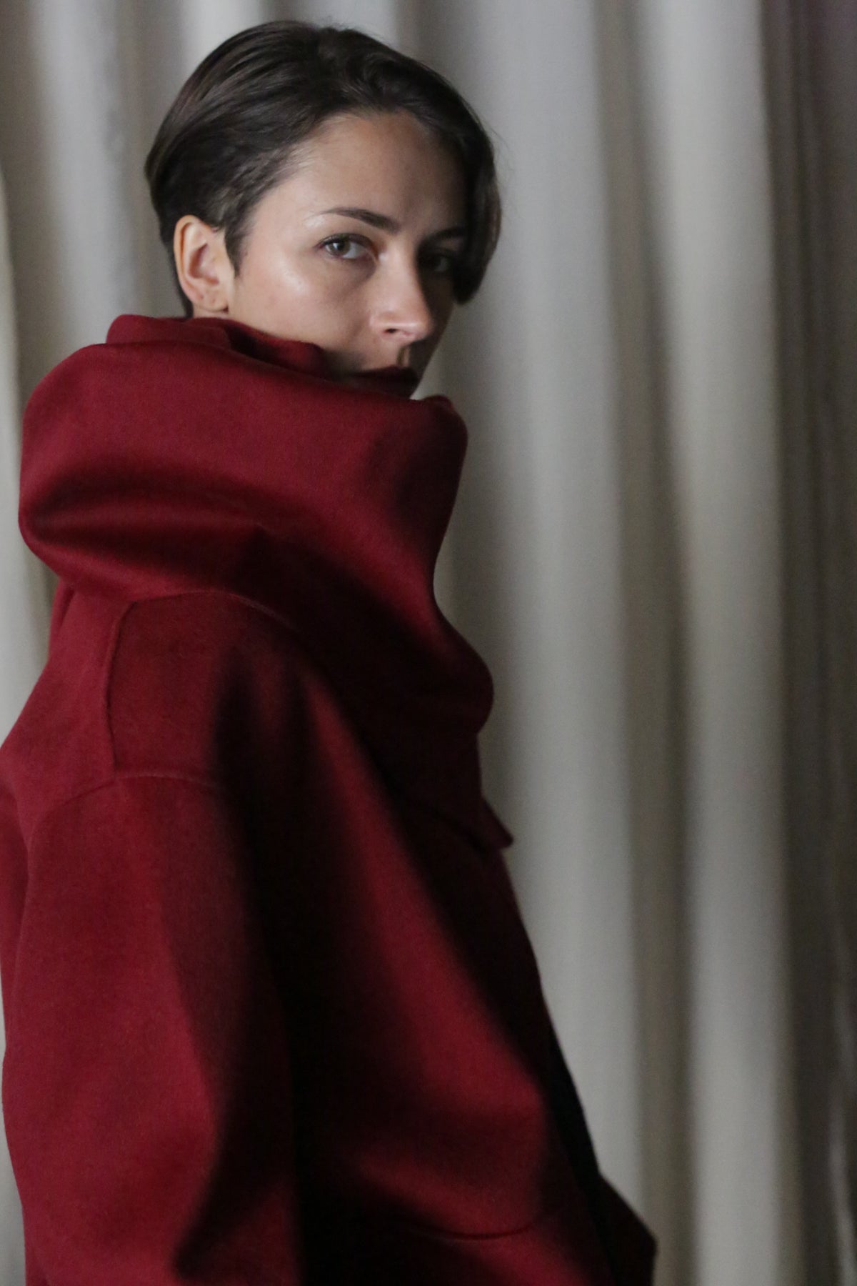 Wearing a high-collared wine X Scarf made from double-faced cashmere, a short-haired person glances over their shoulder in a dimly lit room where light curtains subtly outline the scene.