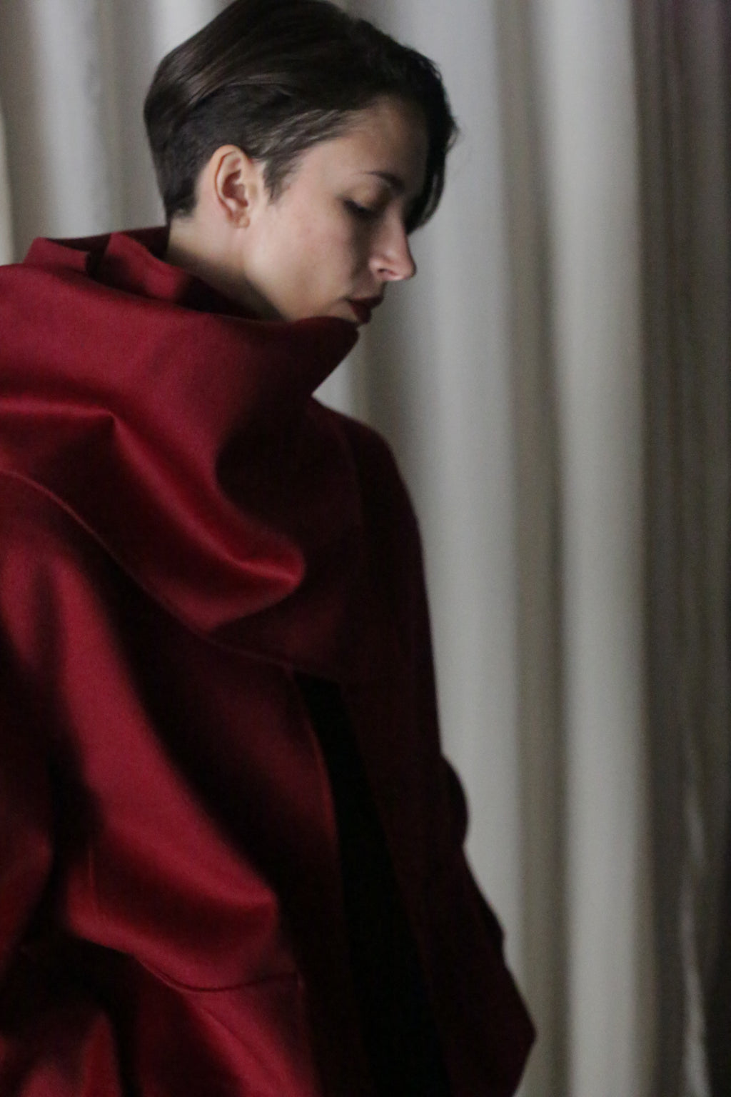 A person with short dark hair wears a large, wine-hued X Scarf draped luxuriously in double-faced cashmere. They stand sideways against a light curtain, highlighting the contrast between their elegant accessory and the bright background.