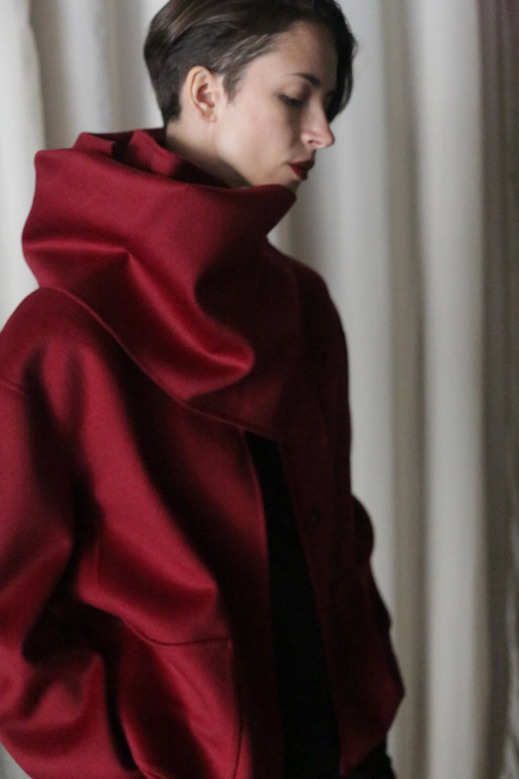 A person with short, dark hair looks downward in a contemplative mood, wearing an oversized high-collared X Scarf made from double-faced wine cashmere, standing before a neutral-colored curtain in soft lighting.