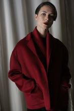 Load image into Gallery viewer, A person with short hair poses in a spacious Bamford Short Double-Face Cashmere coat in a wine hue against a backdrop of softly lit curtains. Their hands rest casually in the coat pockets, maintaining a neutral expression that embodies elements of a contemporary wardrobe.