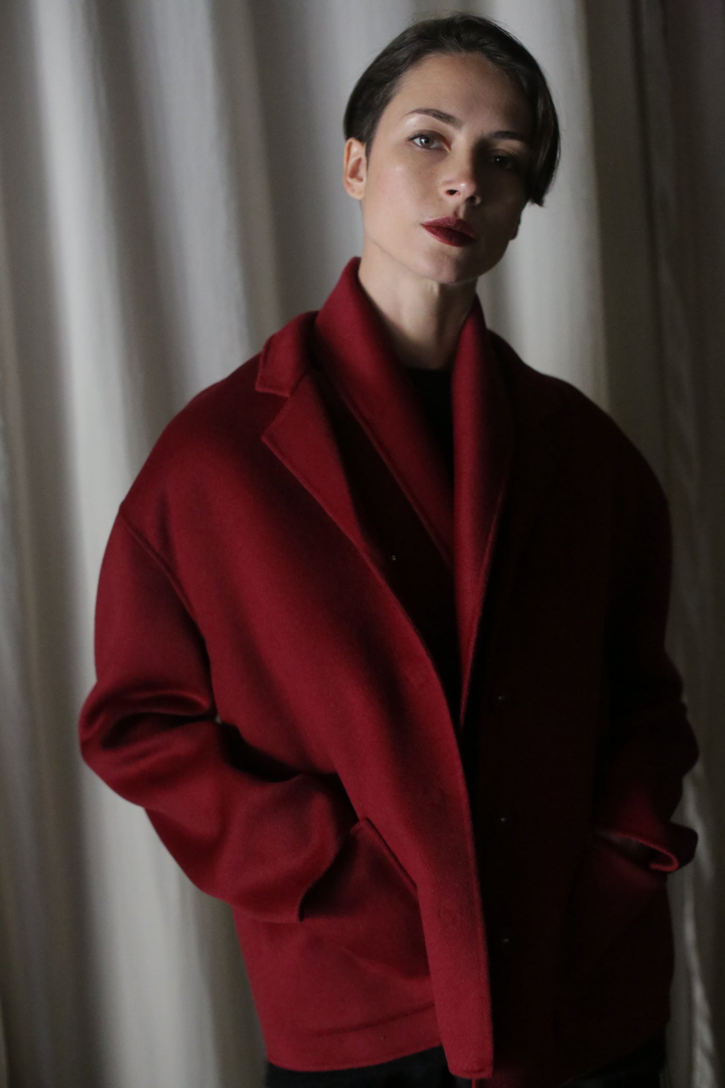 A person with short hair poses in a spacious Bamford Short Double-Face Cashmere coat in a wine hue against a backdrop of softly lit curtains. Their hands rest casually in the coat pockets, maintaining a neutral expression that embodies elements of a contemporary wardrobe.