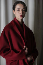 Load image into Gallery viewer, A person with short hair stands in front of a curtain, wearing the Bamford Short Double-Face Cashmere coat in Wine. They have a neutral expression and one hand holds the coat lapel, while soft lighting highlights their face, emphasizing the coat&#39;s modern fit as part of a contemporary wardrobe.