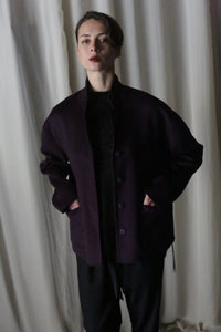 In front of a light-colored curtain backdrop, an individual wears the Bamford Short Double-Face Cashmere jacket in a rich aubergine shade, paired with black pants. With their hands in the jacket's pockets and sporting short dark hair, they exude a contemporary style with modern tailoring.