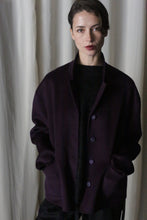 Load image into Gallery viewer, A person with short dark hair stands against a white curtain, showcasing the Bamford Short Double-Face Cashmere in Aubergine from their contemporary wardrobe, with hands in pockets. Their expression is neutral as they look directly at the camera, embodying the essence of modern fit elegance.