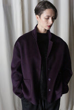 Load image into Gallery viewer, A person with short hair is wearing the Bamford Short Double-Face Cashmere coat in Aubergine over a black outfit, showcasing a contemporary wardrobe. They stand against a light curtain backdrop, looking down with hands in the coat pockets.