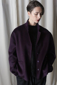 A person with short hair is wearing the Bamford Short Double-Face Cashmere coat in Aubergine over a black outfit, showcasing a contemporary wardrobe. They stand against a light curtain backdrop, looking down with hands in the coat pockets.