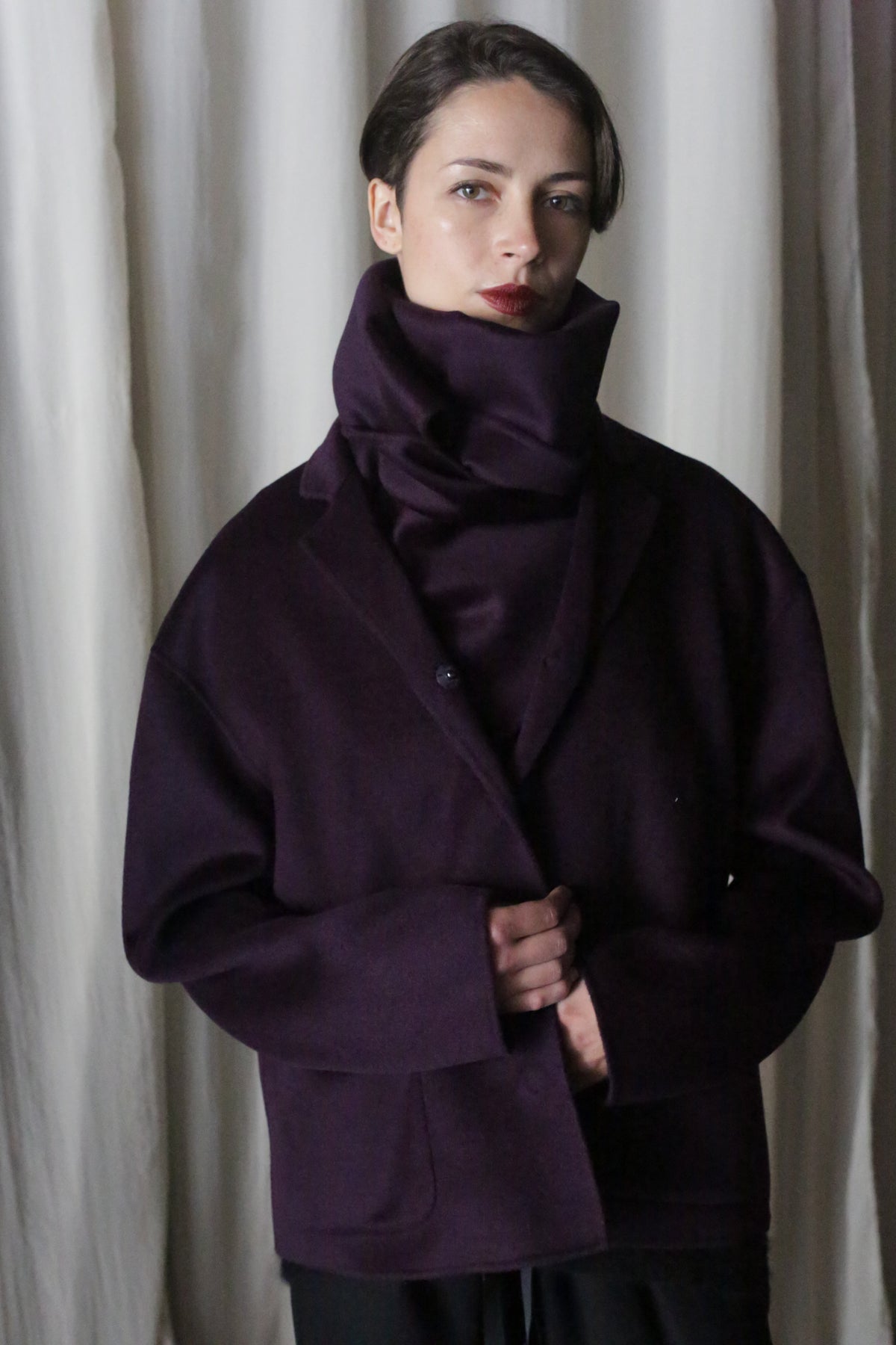 In front of a white, softly lit backdrop, a person with short hair and a neutral expression models an oversized high-collar jacket made from double-faced cashmere in dark aubergine, evoking the elegance and style of the X Scarf - Double-face Cashmere.