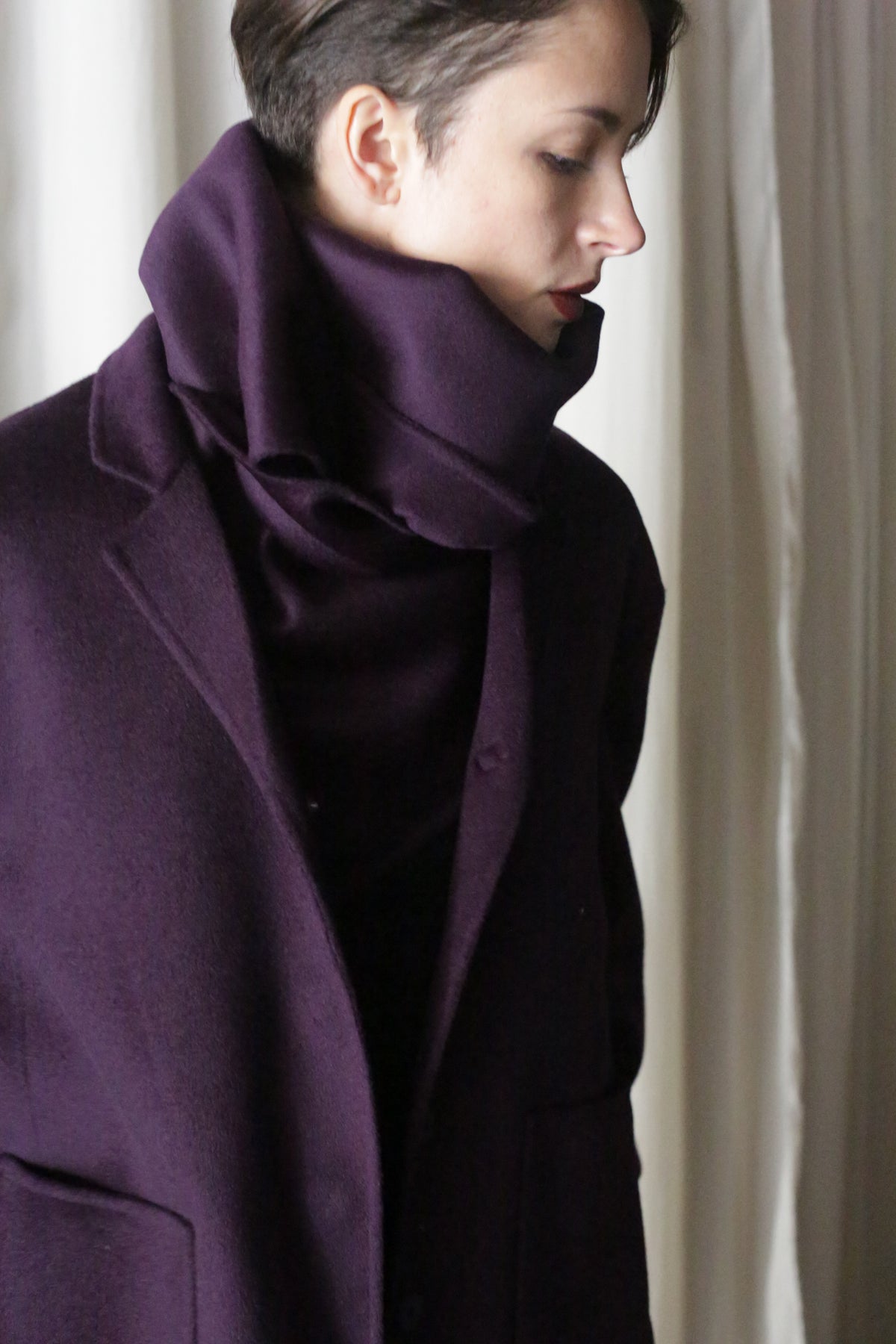 A person wearing an aubergine X Scarf, crafted from double-faced cashmere, with a high collar gazes downward against a backdrop of elegant white curtains.