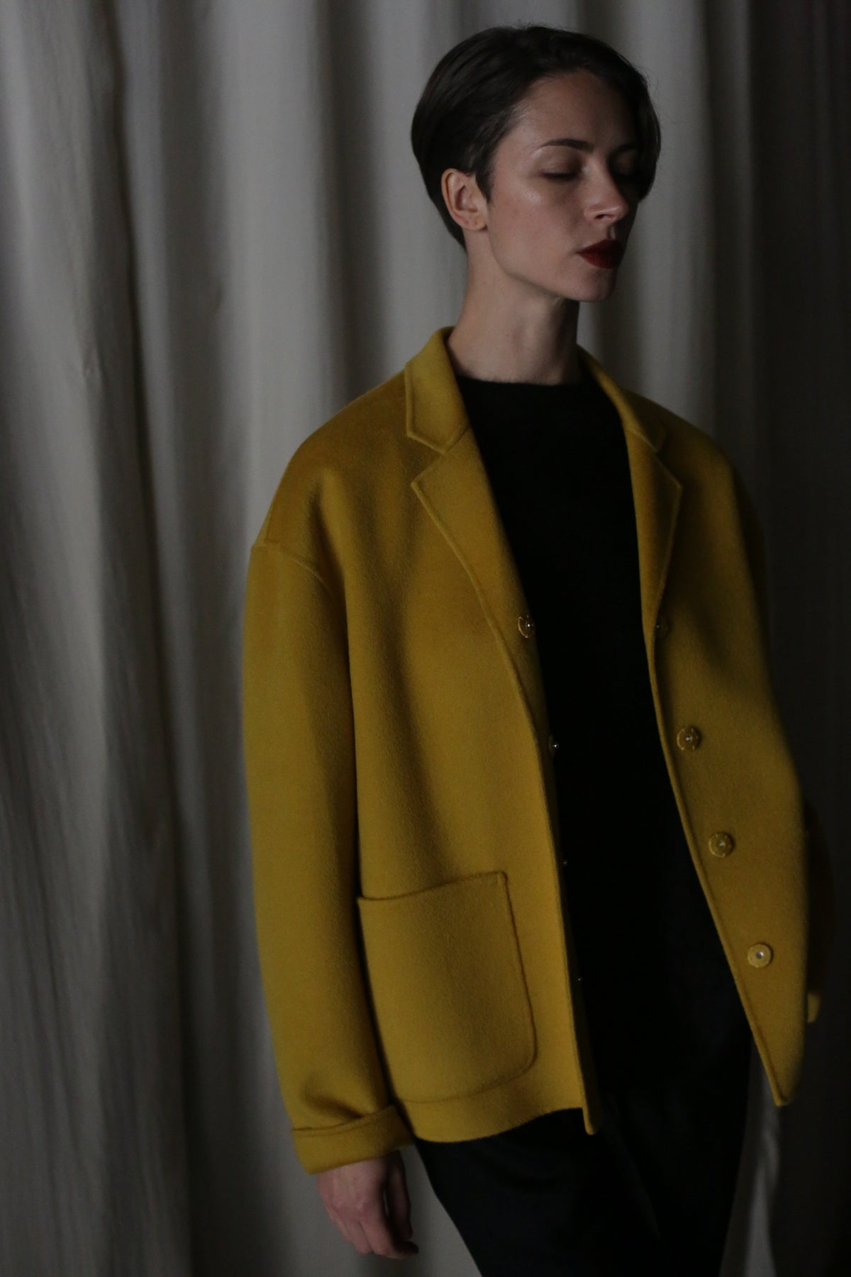 A person with short hair stands with eyes closed, draped in a saffron Bamford Short Double-Face Cashmere coat over a dark outfit—a contemporary wardrobe essential. They are against a softly lit curtain backdrop, embodying understated elegance.