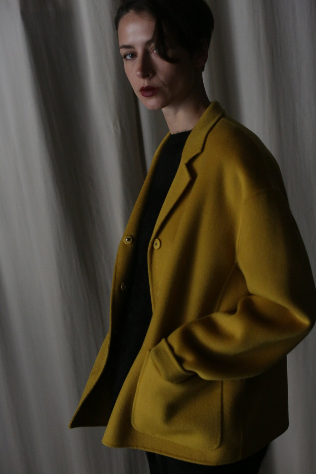 A person in a saffron Bamford Short Double-Face Cashmere coat, a modern wardrobe staple, stands against a neutral curtain backdrop. They have short, dark hair and a serious expression, with one hand tucked in their coat pocket. The soft lighting casts gentle shadows, enhancing the elegance of this ensemble.