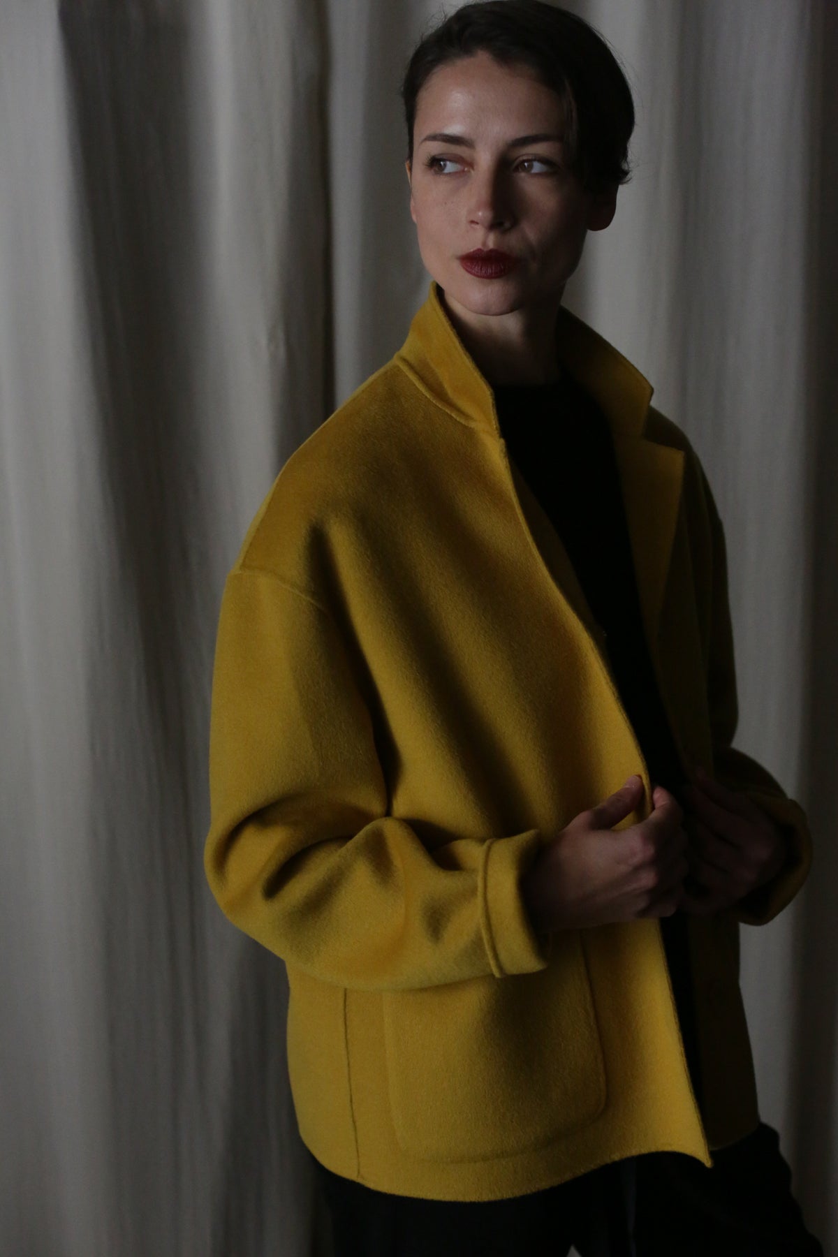 A person stands in front of a curtain, looking to the side. Clad in a Bamford Short Double-Face Cashmere coat in saffron over a black top, they exude modern elegance with their short hair and dark lipstick. The dim lighting casts intriguing shadows across this contemporary wardrobe essential.