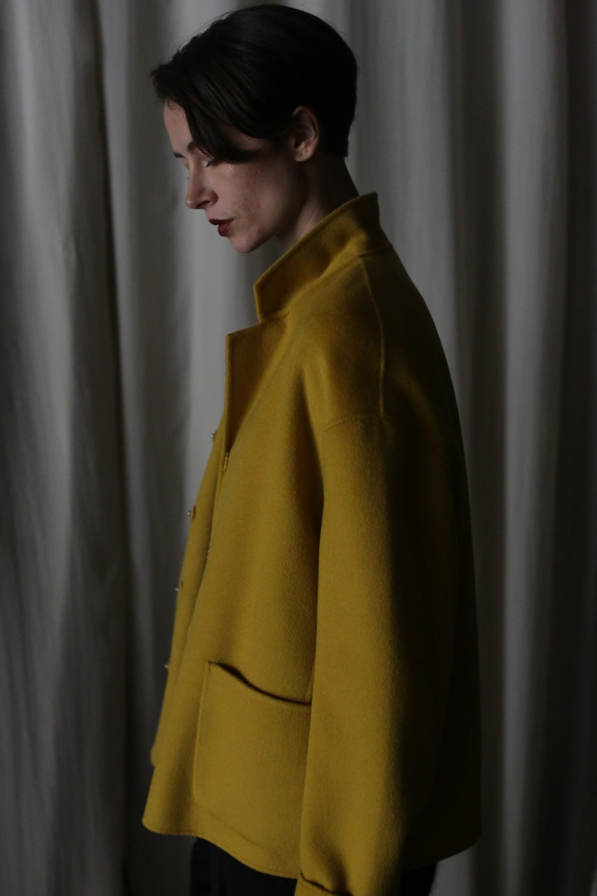 A person wearing a Bamford Short Double-Face Cashmere in saffron stands in profile against a dark, soft curtain backdrop. The dim lighting casts gentle shadows, highlighting the cashmere texture and the person's contemplative expression.