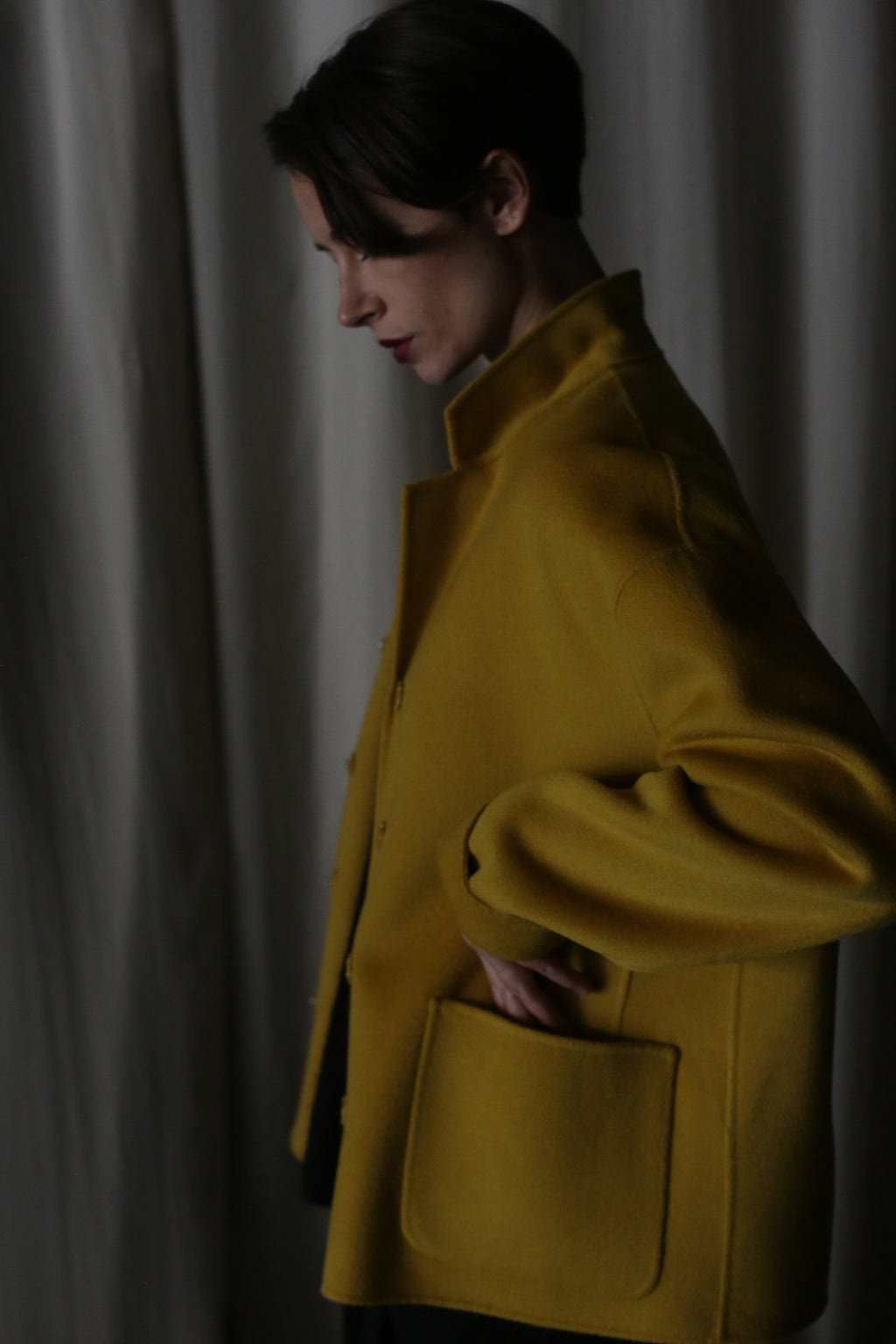 A person stands in profile against a draped curtain, wearing the Bamford Short Double-Face Cashmere in Saffron, a contemporary wardrobe essential. With their hand in the pocket, the dimly lit setting creates a subdued, introspective atmosphere.