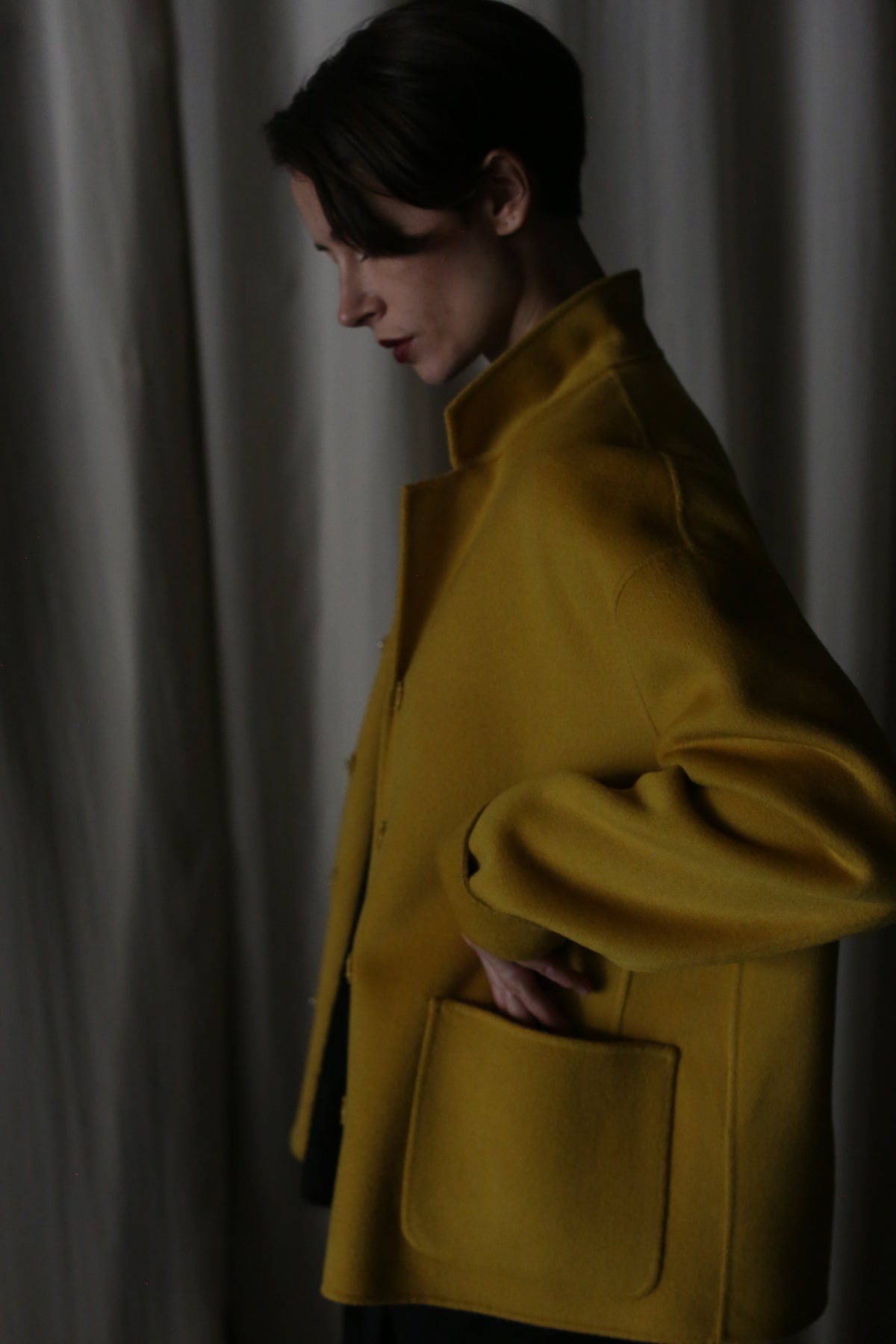 A person stands in profile against a draped curtain, wearing the Bamford Short Double-Face Cashmere in Saffron, a contemporary wardrobe essential. With their hand in the pocket, the dimly lit setting creates a subdued, introspective atmosphere.