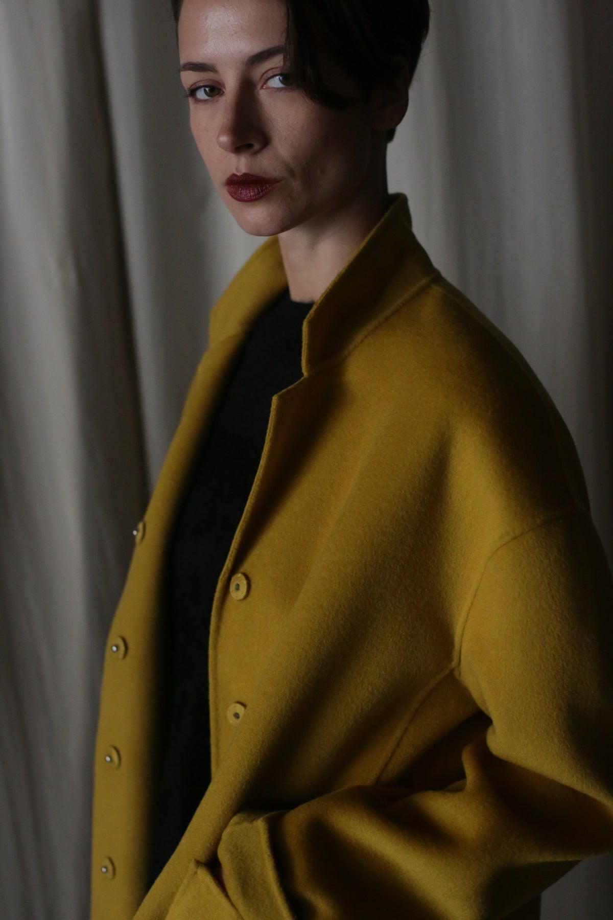 A person with short dark hair stands against a draped curtain background, wearing a Bamford Short Double-Face Cashmere in saffron. The contemporary coat contrasts with their serious expression and the dark shirt underneath.
