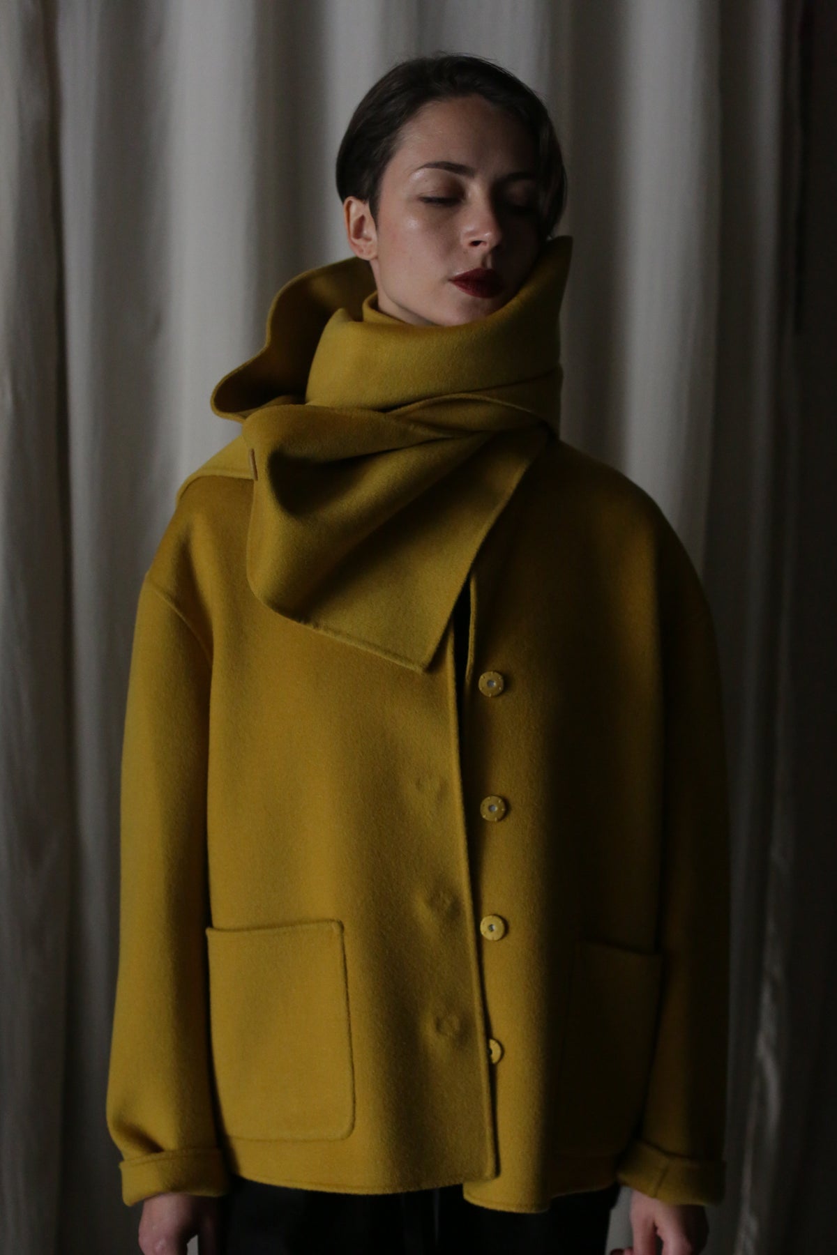 A woman in a saffron coat, a modern wardrobe staple, stands against a neutral background with her eyes closed. The Bamford Short Double-Face Cashmere features a high collar and button-down front, while its large scarf provides both warmth and style.