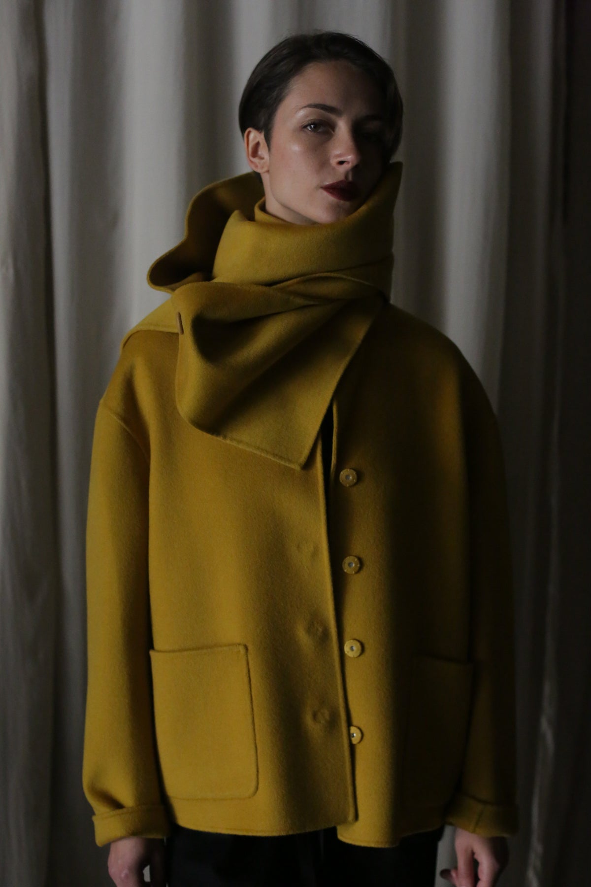 Wearing the Bamford Short Double-Face Cashmere in Saffron, a person is enveloped in a mustard yellow coat with large buttons and an oversized collar that covers their neck and part of their face. This hip-length coat exudes a modern fit against draped white fabric.