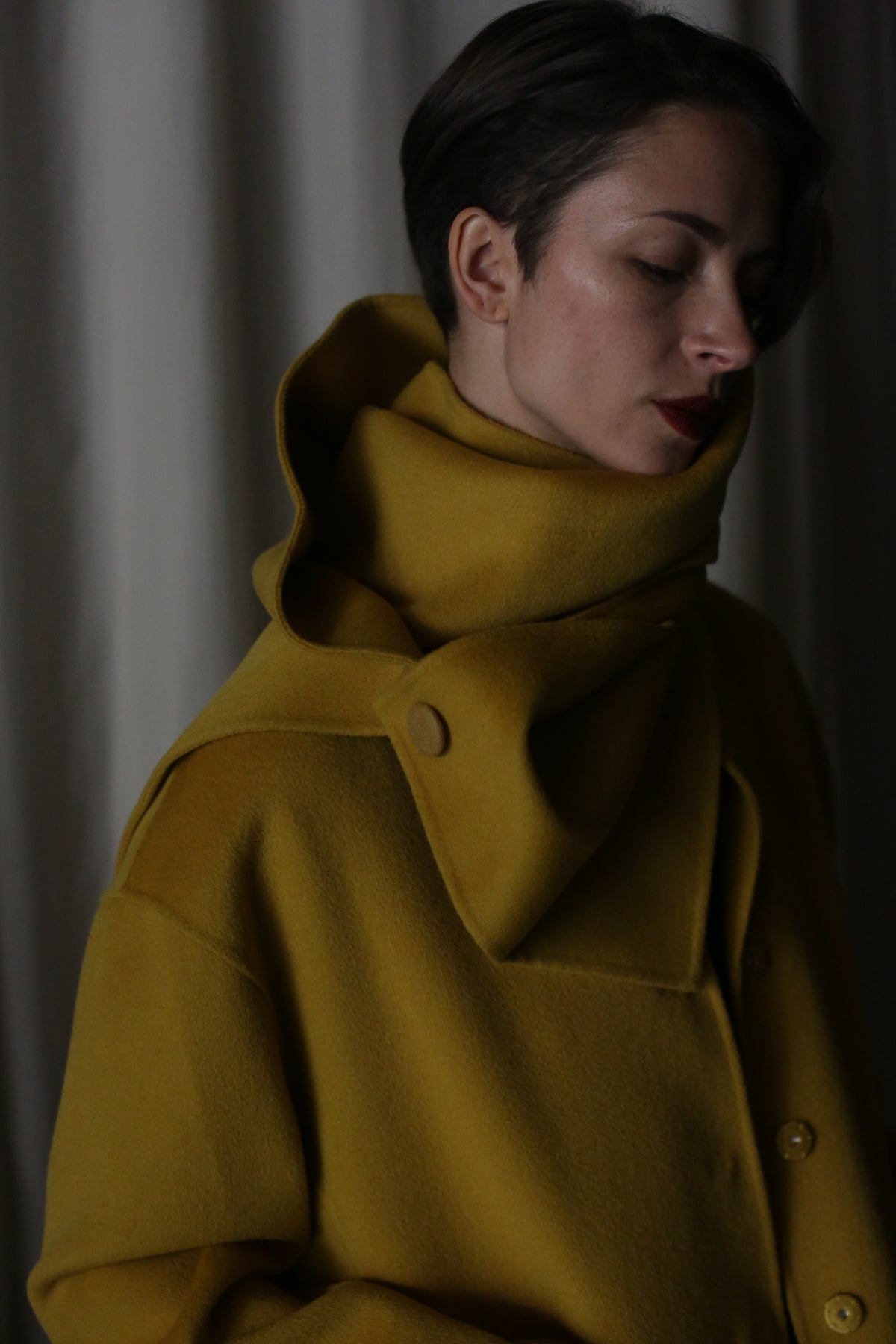 A person with short hair wears a saffron X Scarf made from double-face cashmere, featuring a high collar and buttons. Dim lighting casts shadows on their face, while the softly lit curtain in the background adds an elegant ambiance.