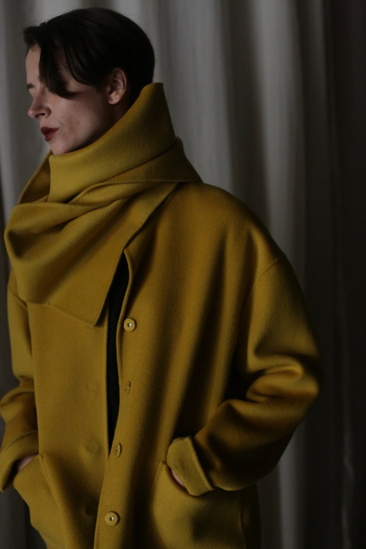 A person with short dark hair stands against a dark curtain, wearing the X Scarf in Saffron—a double-face cashmere coat with a large collar and buttons. They look to their left, hands in pockets.