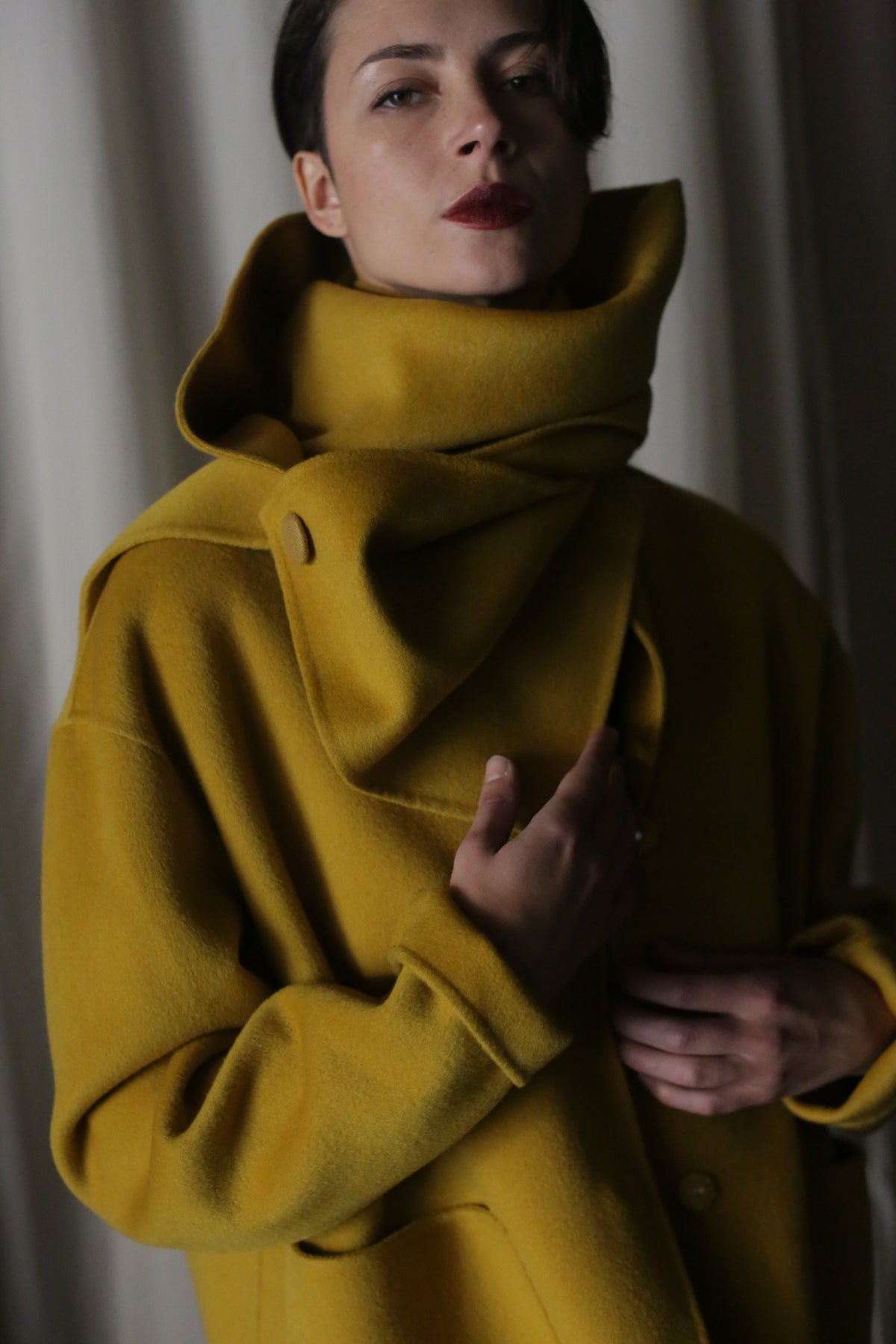 A person models the X Scarf - Double-face Cashmere in Saffron against a neutral background. Their dark hair and red lipstick complement the large-collared coat adorned with grosgrain ribbons. Their hand rests on the coat, and subdued, dramatic lighting adds to the scene's mood.