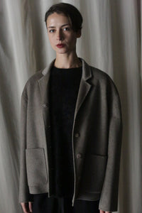 A person with short dark hair wearing the Bamford Short Double-Face Cashmere in Undyed Natural stands in front of a plain background. The luxurious coat features large pockets and buttons. Their expression is neutral, while the soft and dim lighting enhances the elegance of this contemporary wardrobe choice.