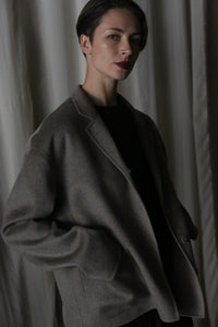 A person with short hair wears the Bamford Short Double-Face Cashmere jacket in a dark, oversized style from a contemporary wardrobe, posing in front of a curtain. The low lighting casts shadows on their face and background, enhancing the modern aura of the scene.