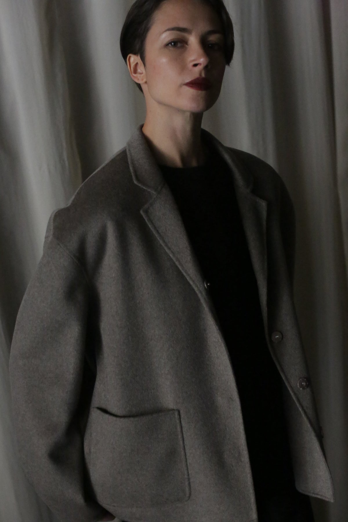 A person with short, dark hair is wearing a modern, dark sweater and the Bamford Short Double-Face Cashmere coat in Undyed Natural. They stand against a backdrop of draped, light-colored fabric, looking slightly upward with a serious expression.