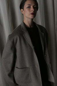 A person with short, dark hair is wearing a modern, dark sweater and the Bamford Short Double-Face Cashmere coat in Undyed Natural. They stand against a backdrop of draped, light-colored fabric, looking slightly upward with a serious expression.