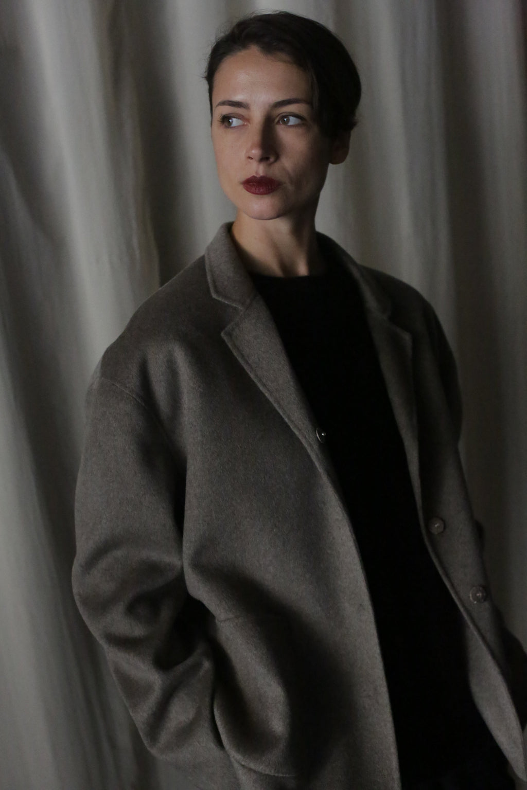 Dressed in a Bamford Short Double-Face Cashmere coat in undyed natural, a person stands against a draped curtain, gazing to the side with a reflective expression. The dim lighting creates gentle shadows, adding depth to the scene and highlighting their contemporary elegance.