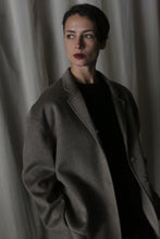 Load image into Gallery viewer, Dressed in a Bamford Short Double-Face Cashmere coat in undyed natural, a person stands against a draped curtain, gazing to the side with a reflective expression. The dim lighting creates gentle shadows, adding depth to the scene and highlighting their contemporary elegance.