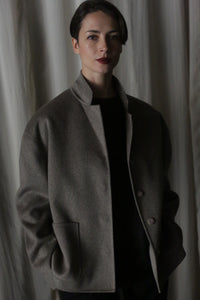 A person with short hair stands against a backdrop of draped curtains, wearing the Bamford Short Double-Face Cashmere in Undyed Natural from their contemporary wardrobe. Their composed expression adds to the modern elegance of the scene.